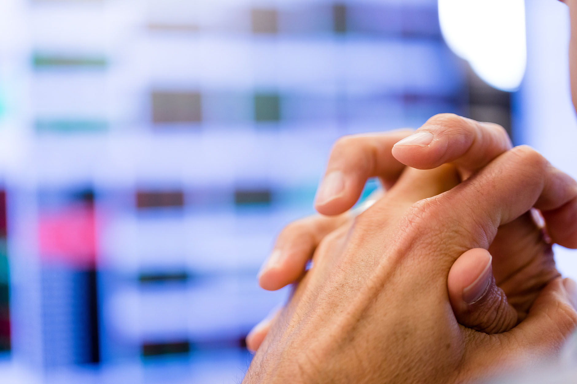 A close view of two hands clasped with a blurred computer display in the background. 