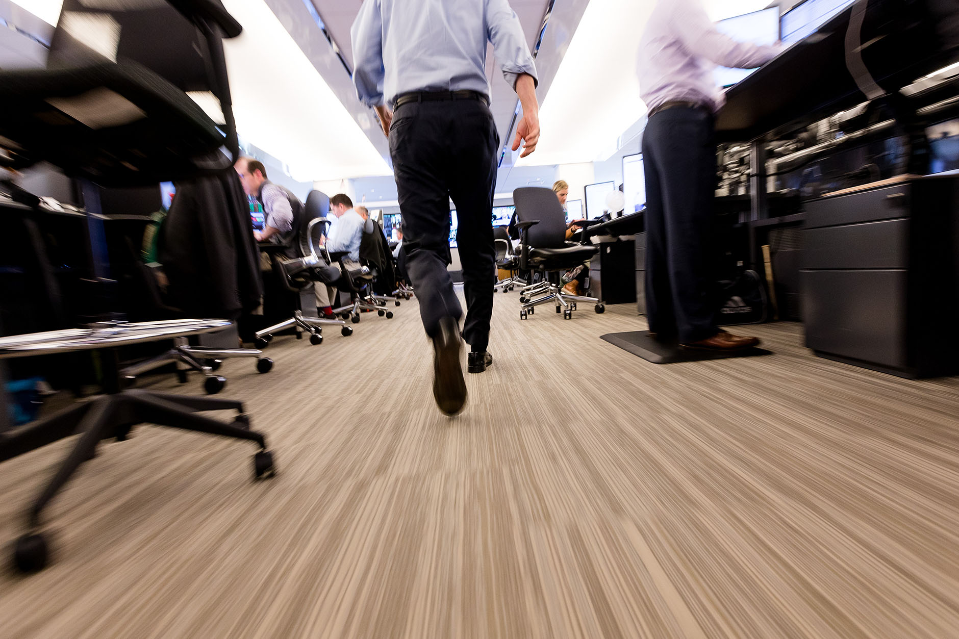 A low angle view of a figure walking through a crowded and brightly lit office. 