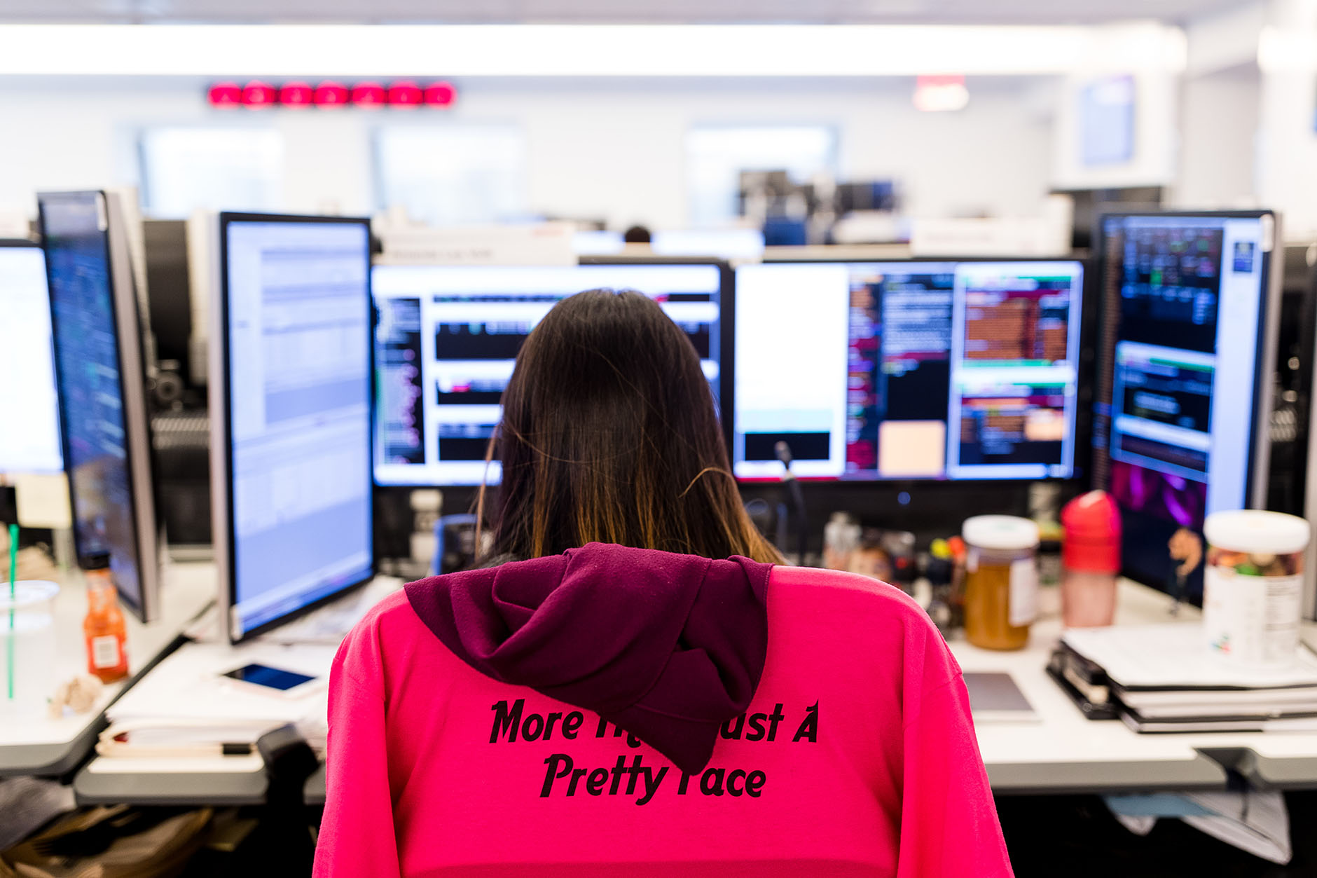 A view from behind of a trader at her workstation. A red sweatshirt draped over the chair is printed with the text: More than just a pretty face.