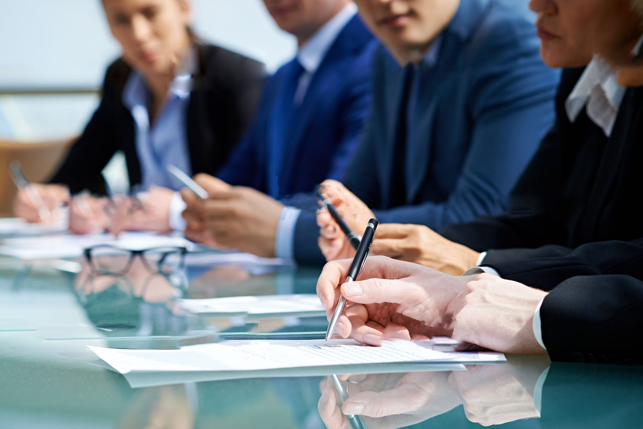 A close up someone taking notes at a long glass-topped conference table with several other people in suits seated in the background. 